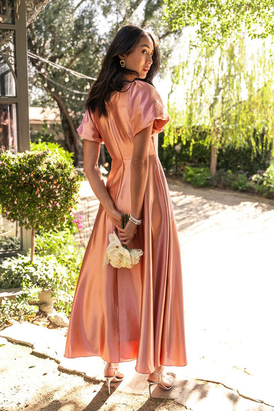 Wearing the Tessie Dress in Apricot Crush by Ivy City Co, a woman stands outdoors on a sunny day, holding white flowers and looking over her shoulder. Green trees and bushes provide a lush backdrop.
