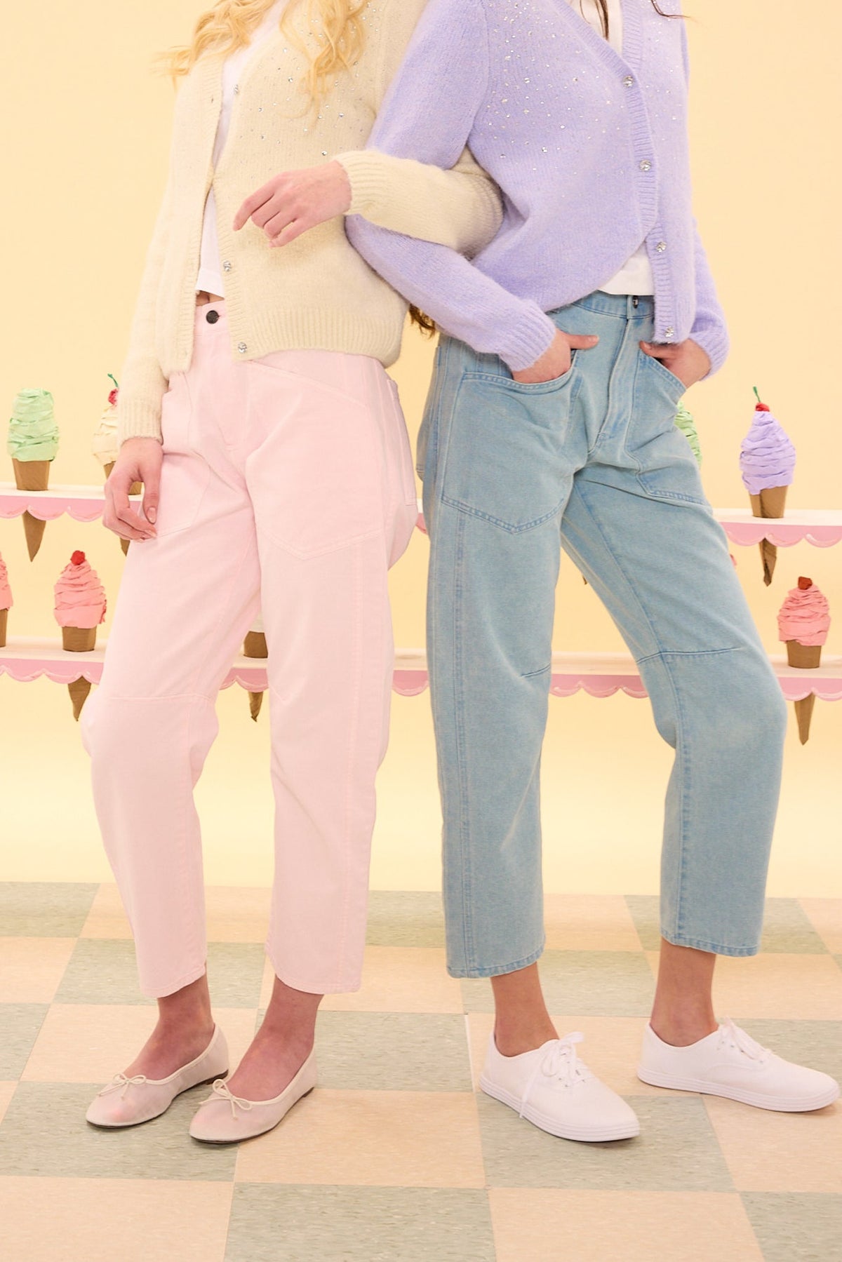 Two women stand side by side, arms linked. One wears the Amanda Pants in Light Pink by Ivy City Co paired with a cream sweater; the other pairs high rise denim pants with a lavender cardigan. Ice cream cones decorate the pastel background.