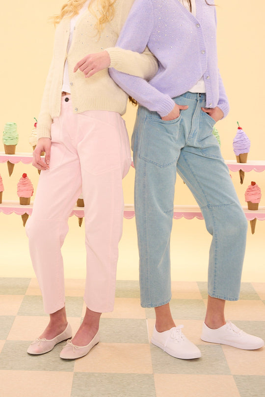 Two women stand side by side in pastel outfits—one wears the Amanda Pants in Light Pink from Ivy City Co paired with a cream cardigan, while the other sports high-rise denim pants and a lavender sweater. Ice cream cones are displayed on a pastel counter behind them.