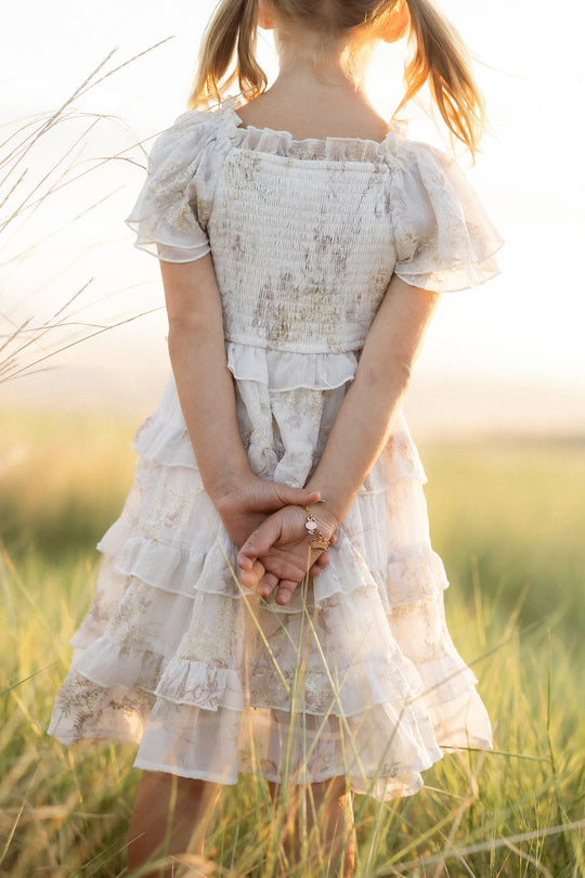 A young girl wearing the Ivy City Co Mini Winnie Dress in Bright White Wispy Wildflower stands in tall grass, facing away from the camera with her hands clasped behind her back as sunlight creates a soft, warm glow around her.