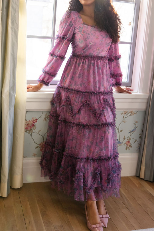 A woman stands by a window in the Catherine Dress in Climbing Vine Bloom from Ivy City Co, its long, ruffled silhouette and puffed sleeves catching the sunlight as it streams onto the wooden floor. She wears pink heels to complete her look.