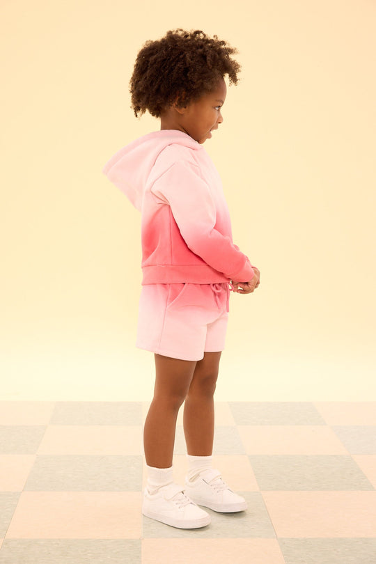 A young child stands sideways on a checkered floor against a plain light yellow background, wearing the Ivy City Co Mini Ash Hoodie in Pink Ombre paired with matching pink ombre girls’ sweatshorts, white socks, and white sneakers.