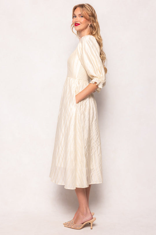 A woman with long blonde hair is smiling as she stands sideways with her hands in the pockets of the Ivy City Co Tindra Dress in Cream Sequin, a nursing-friendly midi dress featuring puff sleeves. She pairs it with beige heeled sandals.