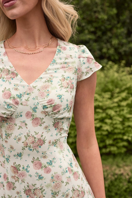 A woman stands outdoors, wearing the Ivy City Co “Fiona Dress in Countryside Peony Blossom,” which features pink and green floral patterns on a white background. Only her upper body is visible; she has wavy blonde hair and a delicate gold necklace, with green foliage behind her.