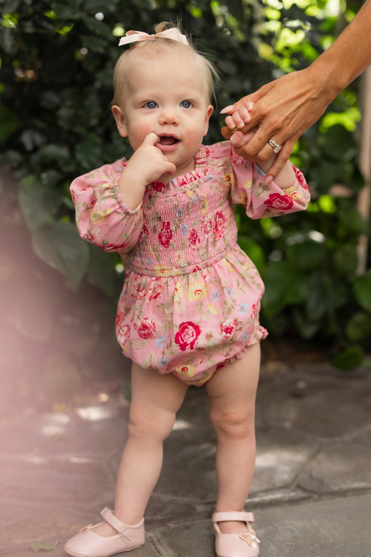 A baby with light hair, wearing a bow and matching pink shoes, stands on a stone path surrounded by lush greenery. She is dressed in the Ivy City Co Baby Madeline Romper in Pink Vintage Poppies, featuring a smocked bodice and charming floral print, while holding an adult’s hand and touching her mouth.