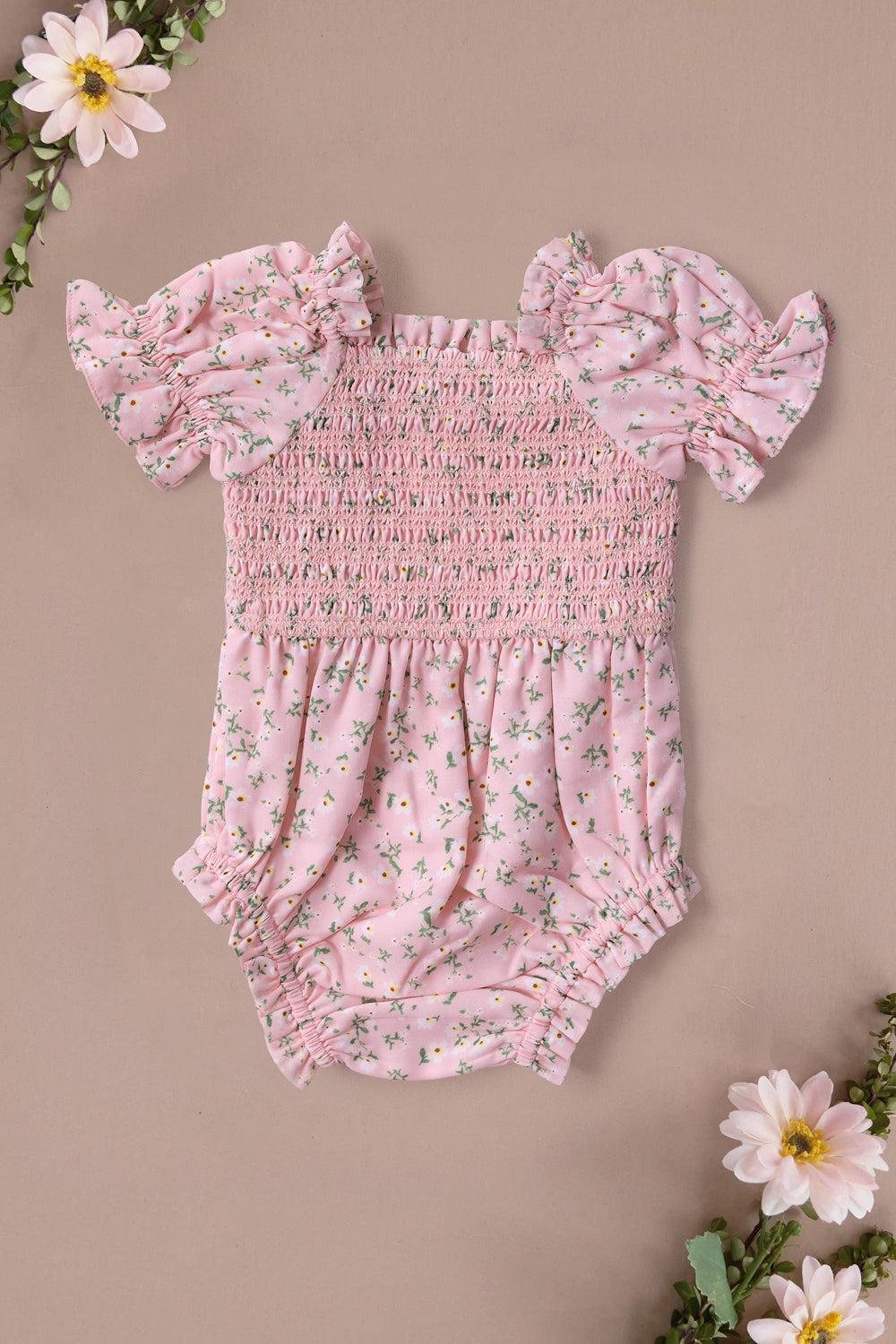 The Ivy City Co Baby Valencia Romper in Pink Ditsy Floral features ruffled sleeves and a smocked bodice, adorned with delightful white and green ditsy floral prints on a soft pink background. Surrounded by pink and white flowers on beige, this charming romper offers an adorable and timeless look for your little one.