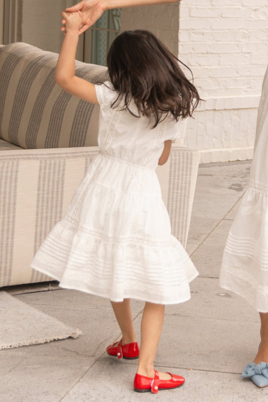 A young girl in the Ivy City Co Mini Palermo Dress in White, paired with red shoes, spins while holding an adult’s hand near a striped sofa and white brick wall.