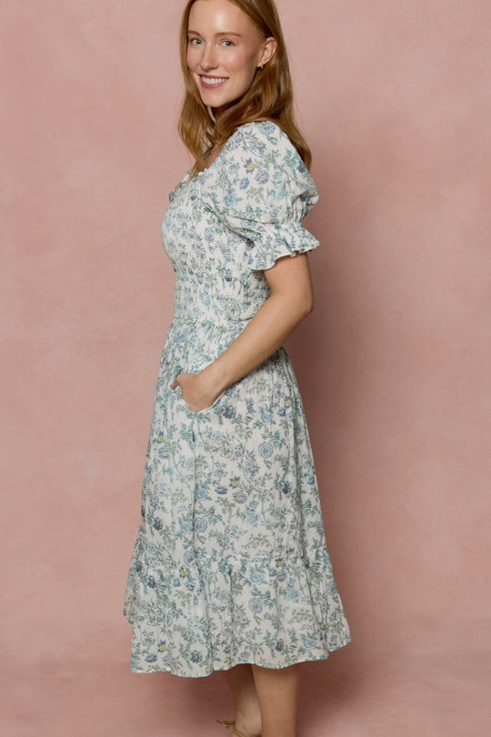 A woman stands sideways, smiling, in the Ivy City Co Valencia Dress in Ivory Climbing Vine Bloom—a maternity-friendly cotton midi dress with short puff sleeves and pockets—set against a soft pink background.