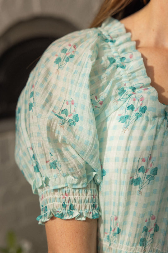 A close-up of a woman’s shoulder wearing the Ivy City Co Jessie Dress in Bloom Gingham, featuring a light blue gingham with floral print, puff sleeves, and ruffled trim—this nursing-friendly design is effortlessly stylish.