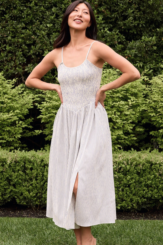 A woman stands smiling in a garden, wearing the Ivy City Co Maxwell Dress in Blue & White Pinstripe—a light-colored, sleeveless midi dress with thin straps and a front slit. This stylish and practical piece is perfect as a nursing-friendly or maternity dress. Green bushes and trees provide a lush backdrop.