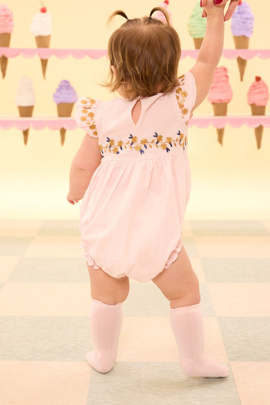 A toddler wearing the Ivy City Co Baby Hattie Romper in Embroidered Spring Floral and knee socks stands on a pastel-tiled floor, reaching up with her back to the camera. The pale yellow background is decorated with colorful ice cream cones, while the romper’s delicate embroidered spring floral details create a charming touch.