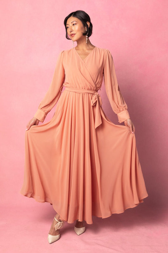 A woman wearing the Andie Dress in Apricot Crush by Ivy City Co poses against a pink background, holding out the long maternity maxi skirt. She has dark hair styled in an updo, and accessorizes with earrings and light heels.