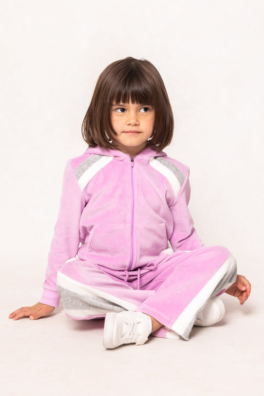 A young girl with straight brown hair sits cross-legged against a plain white background, wearing the Ivy City Co Mini Kailey Zip Up Velour Sweatshirt in Pink paired with gray track pants and white sneakers. She gazes slightly to the side with a neutral expression.