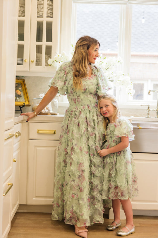 A woman and a young girl, both wearing the Ivy City Co Florentina Dress in Pistachio Aquarelle Fleur, stand smiling together in a bright, elegant kitchen with white cabinets and gold accents.