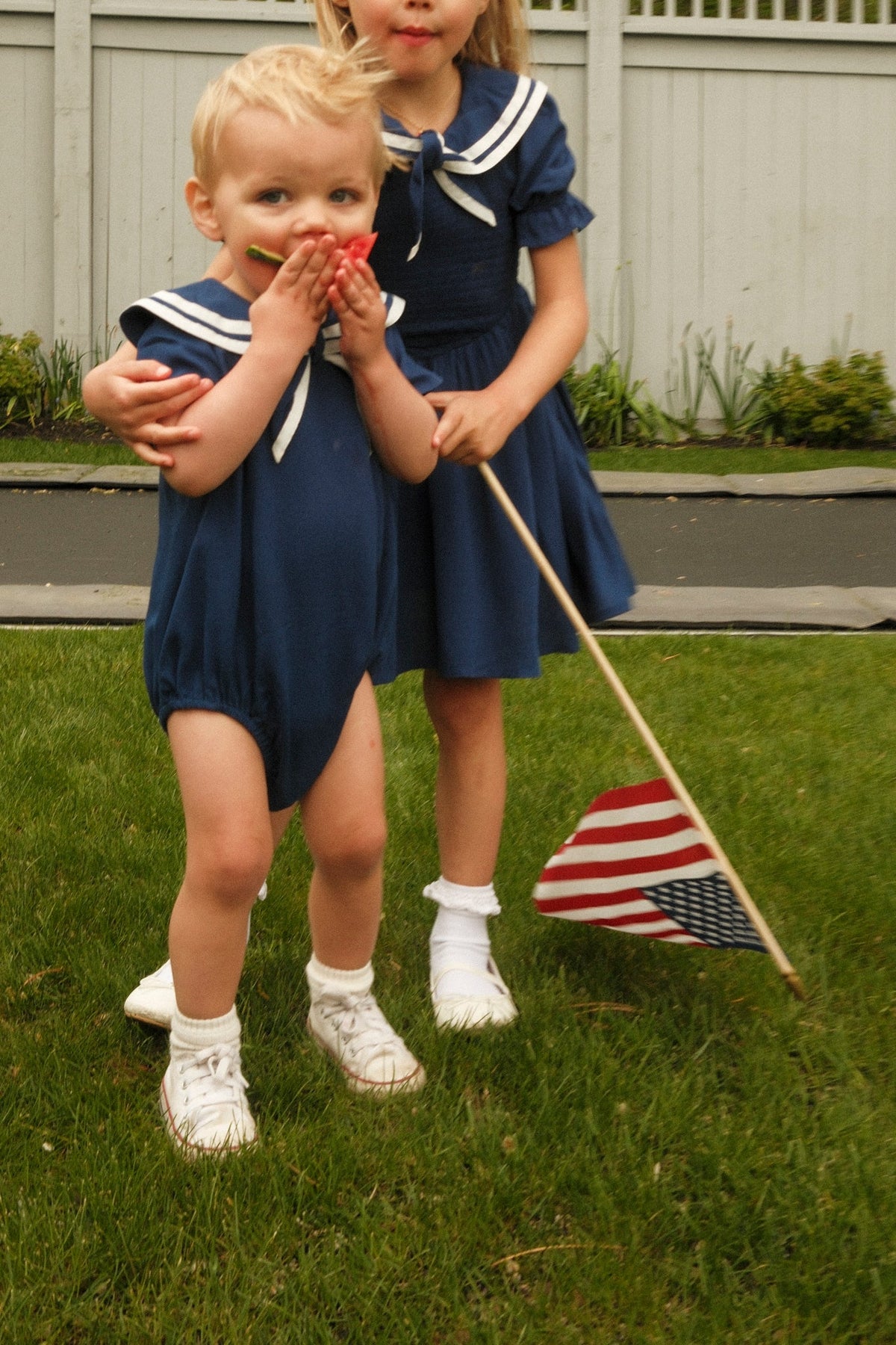 Two young children dressed in matching Ivy City Co Baby Sailor Rompers in Navy stand on the grass. One covers their mouth while the other holds an American flag, with a white fence and greenery visible in the background.