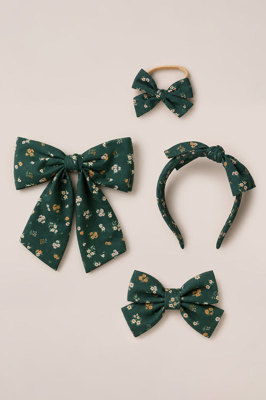 Four green cotton hair accessories from Ivy City Co, called “Hair Bows in Evergreen Lovelorn Ditsy,” each featuring small white and yellow floral patterns: a large hair bow, a headband with a bow, a small bow on an elastic band, and a medium-sized bow. All are displayed together on a neutral background.