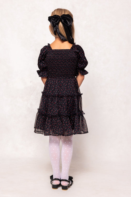 A young girl stands facing away, wearing the Ivy City Co Mini Madeline Dress in Black Ditsy Floral, paired with white polka dot tights, black shoes adorned with bows, and a large black ribbon in her hair.