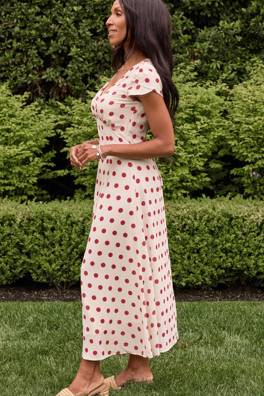 A woman strolls across the grass beside neatly trimmed green bushes, her dark brown hair loose and beige sandals on her feet. She wears the Fiona Dress in Red Polka Dot by Ivy City Co—a cream-colored, midi-length summer dress adorned with red polka dots that lends an elegant touch to the breezy scene.