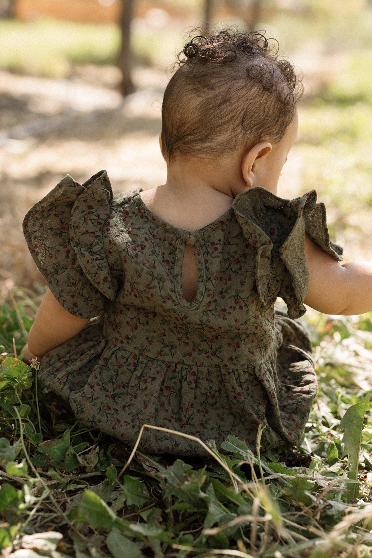 A baby with short curly hair sits on green grass, wearing the Ivy City Co Baby Hattie Romper in Woodland Floral, featuring flutter sleeves. The baby is facing away, with sunlight and trees softly blurred in the background.