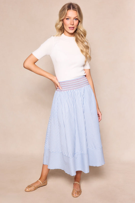 A woman with long blonde hair stands against a beige background wearing a white short-sleeve top and the Ivy City Co Bluebell Skirt in Blue & White Stripes, which features a smocked waistband. She completes the look with tan flats.