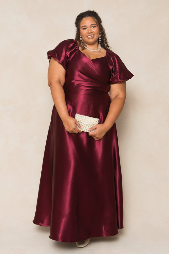 A woman stands smiling against a plain light beige background, wearing the Ivy City Co Tessie Dress in Wine Satin with puff sleeves. She holds a pearl clutch and accessorizes with pearl earrings and a necklace, her curly hair styled down.