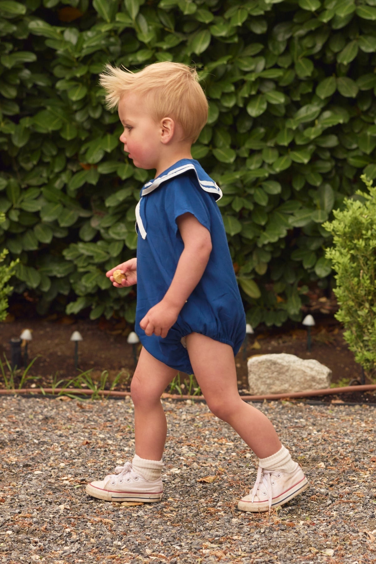A young child wears the Ivy City Co Baby Sailor Romper in Navy with white sneakers while walking outside on a gravel path, holding a small object and surrounded by green bushes.