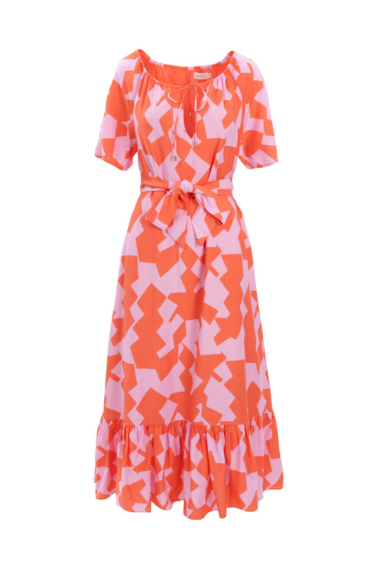 The Haven Dress in Geometric by Ivy City Co is a short-sleeve, knee-length dress featuring a red and pink geometric pattern, a waist tie, and a ruffled hem. It has a scoop neckline adorned with a small bow at the front.