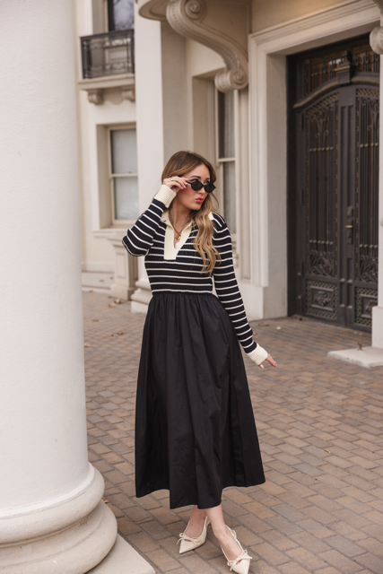 A woman wearing sunglasses and the Adelina Dress in Black by Ivy City Co—with its black and white striped knit bodice and black skirt—stands on a paved walkway near white columns and an ornate building entrance, looking to the side.
