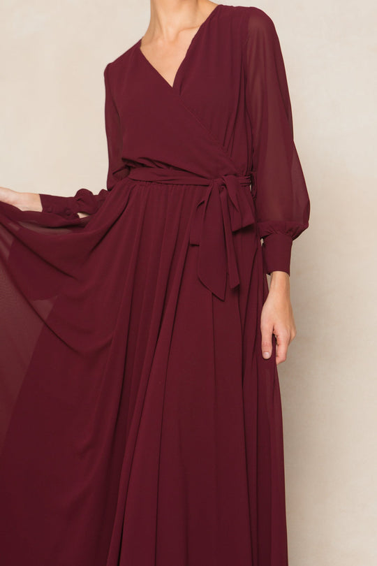 A person wearing the Ivy City Co Andie Dress in Wine Chiffon, featuring long sleeves and a tied waist sash, holds out part of the flowing skirt against a neutral background. The face is not visible.