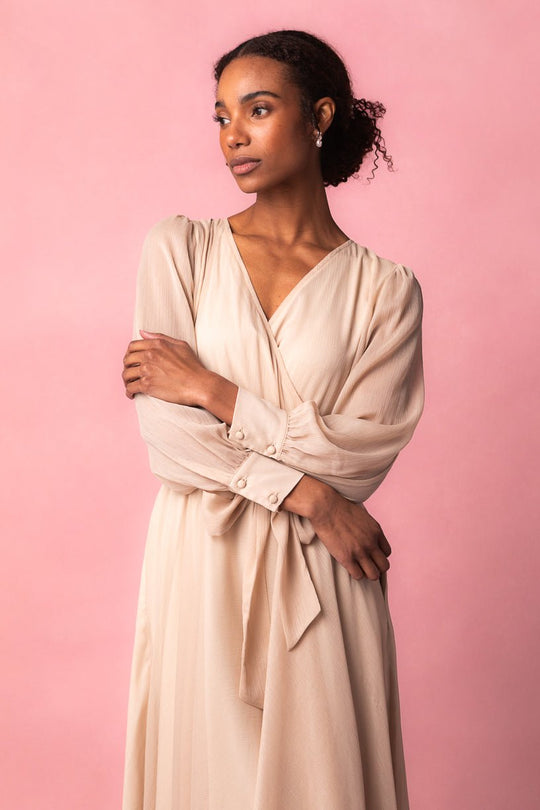 A woman wearing the Andie Dress in Champagne by Ivy City Co stands against a pink background, looking to the side with her arms gently crossed and an elegant, composed expression.