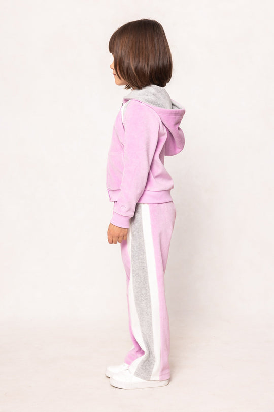 A young child with brown hair stands in profile, wearing the Ivy City Co Mini Kailey Zip Up Velour Sweatshirt in Pink, paired with matching pants featuring a gray and white stripe down the sides and white shoes, against a plain light background.