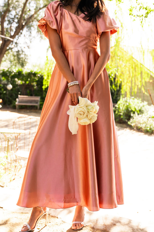 A woman stands outdoors in sunlight wearing the Ivy City Co Tessie Dress in Apricot Crush, styled with strappy heels and bracelets. She holds a small bouquet of white roses, with greenery and a bench visible in the background.