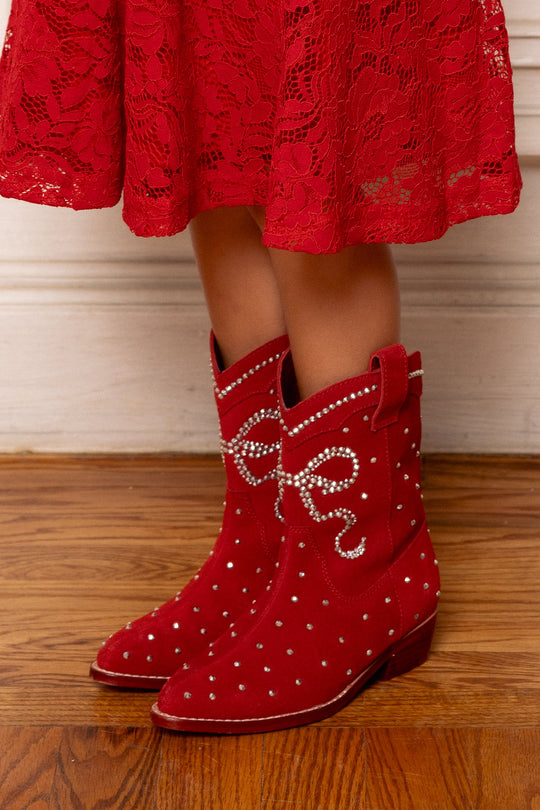 A child wearing a red lace dress and Ivy City Co’s Mini Channing Bootie in Red, adorned with white pearls, stands on a wooden floor.