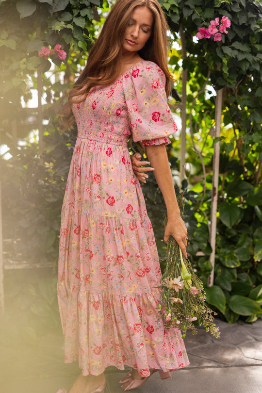 A woman in a long, pink floral maxi dress stands in a lush garden holding a bouquet of wildflowers, surrounded by green leaves and pink blossoms. She wears the Ivy City Co Madeline Maxi Dress in Pink Vintage Poppies—a nursing-friendly style that brings elegance and comfort to the serene scene.