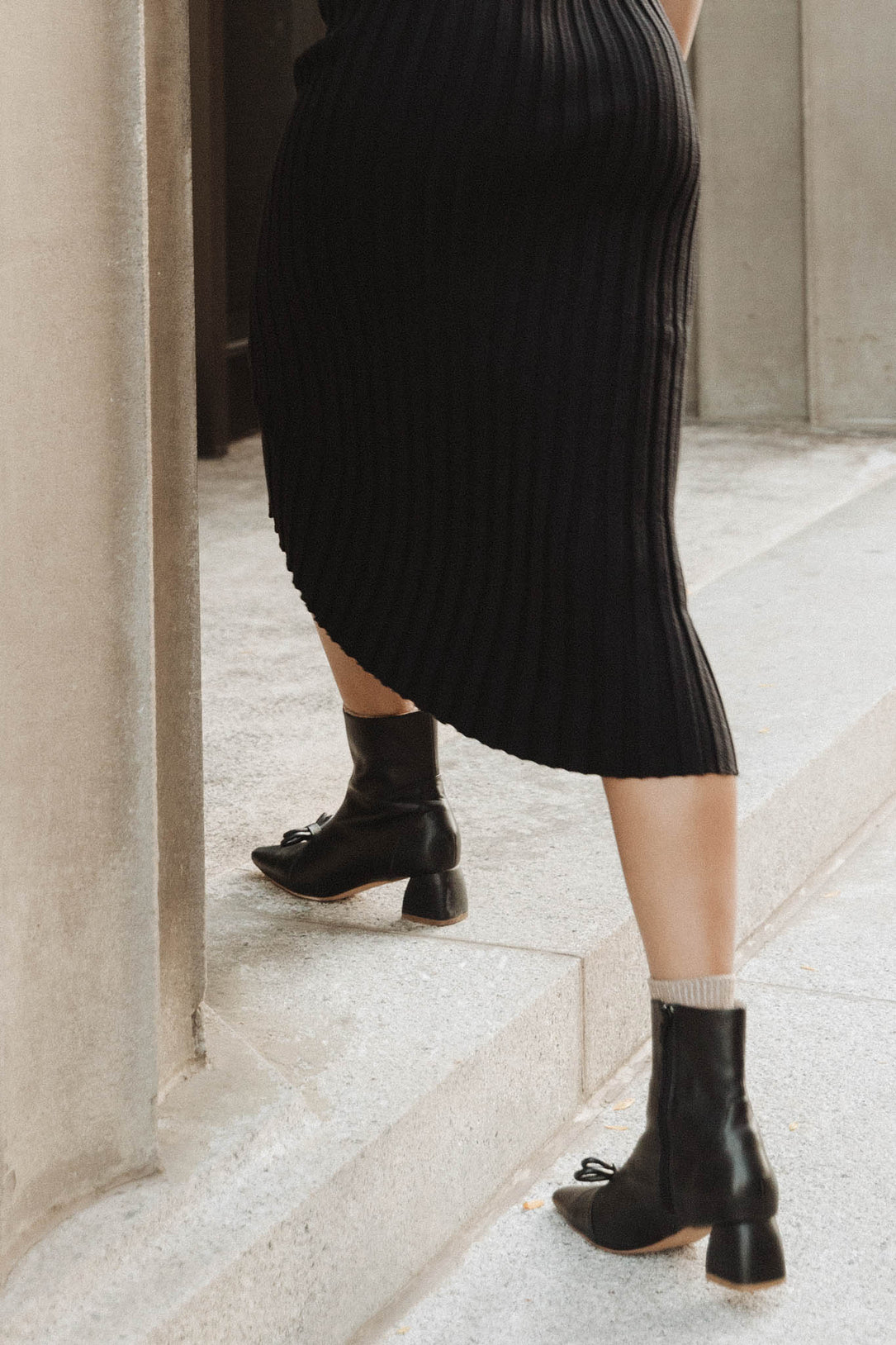 Wearing a black ribbed skirt and light socks, a person in Ivy City Cos Ivy Bow Ankle Boot—black leather, low heel, and subtle bow detail—walks up concrete steps beside a stone wall.