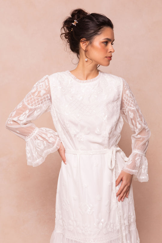 A woman in the Ivy City Co Queenie Dress in White—a long-sleeved lace gown with embroidered patterns and hand-embellished sequins—stands against a beige background, gazing to the side with one hand on her hip.