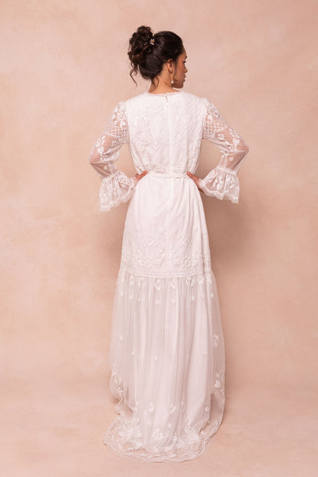 A woman faces away from the camera in the Ivy City Co Queenie Dress in White, a long lace gown with sheer sleeves and hand-embellished sequins. Her hair is elegantly styled, set against a soft beige background.