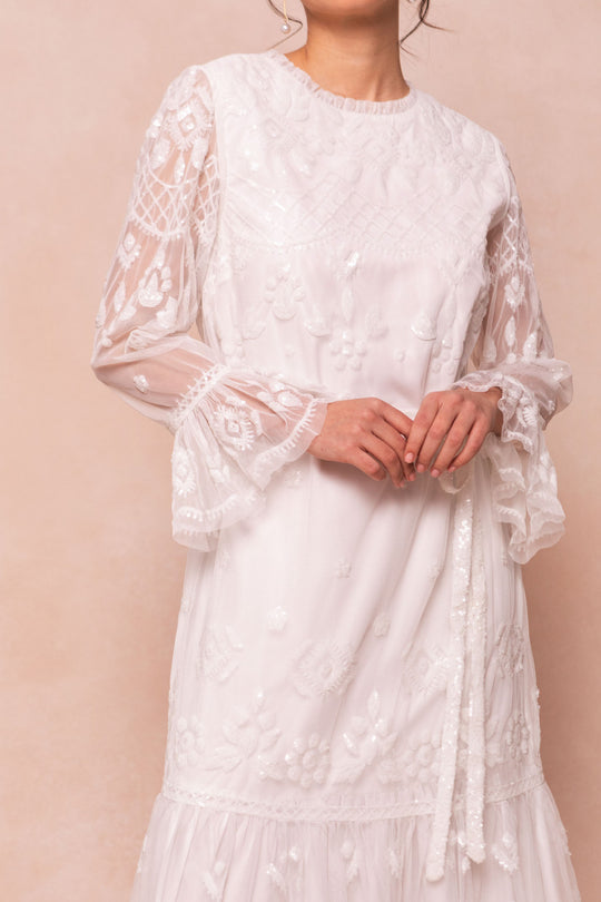 A woman in the Ivy City Co Queenie Dress in White—a tulle gown with sheer, embroidered sleeves and floral details—stands against a soft beige background. Her face is partially out of frame.