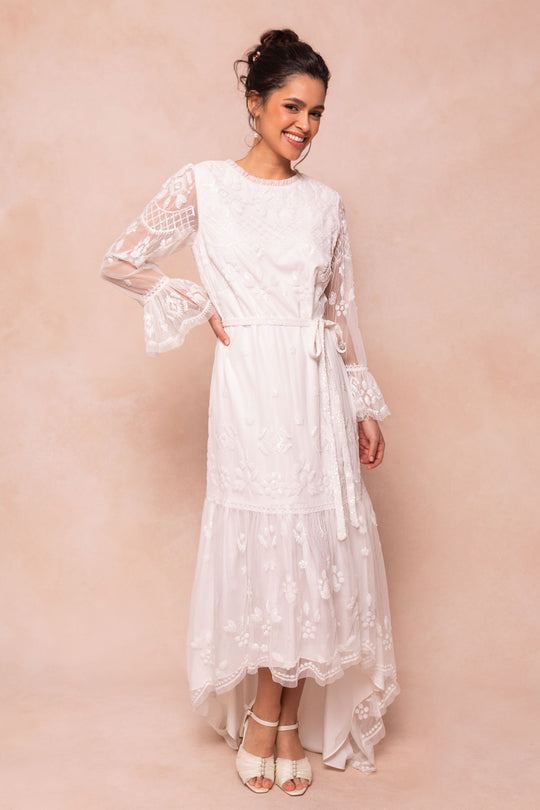 A woman smiles against a soft beige background wearing the Ivy City Co Queenie Dress in White, featuring long sheer sleeves, floral embroidery, hand-embellished sequins, a tie waist, and high-low hem. She pairs it with white heels and an updo.