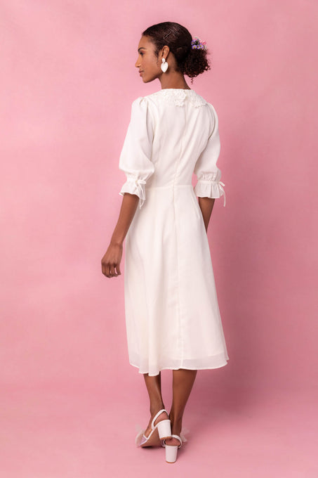 A woman standing in a pink backdrop wearing a cream-colored knee-length dress with a round neckline, puff sleeves, non-functional buttons, and a lace peter collar detail. She is also wearing earrings and shoes.