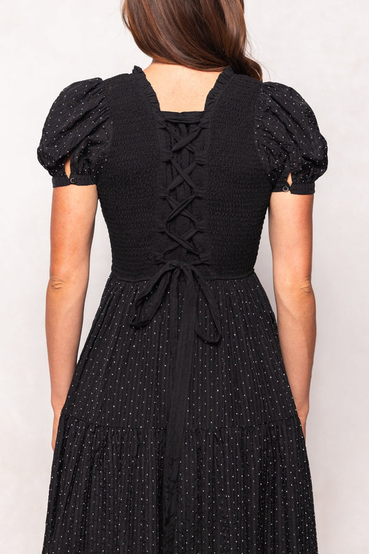 A woman stands facing away from the camera, wearing the Ivy City Co Darby Dress in Black Diamonte—a black tiered midi dress with puff sleeves, a smocked back, lace-up corset detail, and a subtle dotted pattern. The design is maternity friendly.