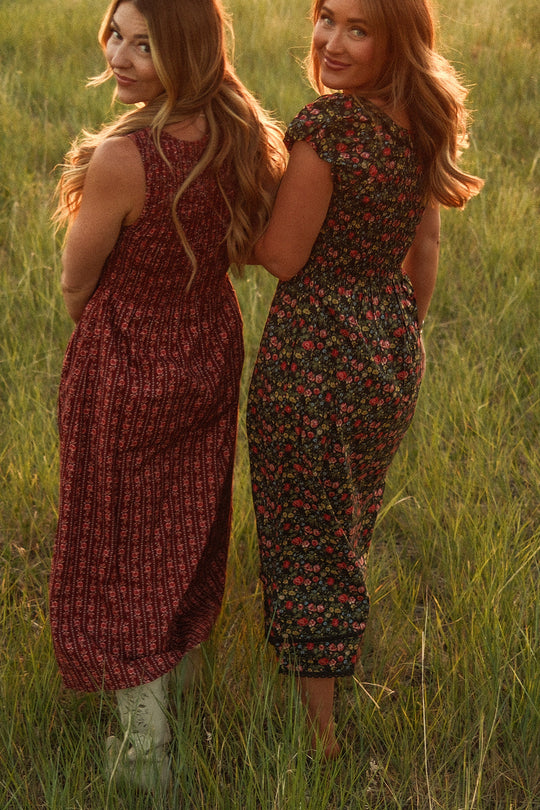 Two women with long wavy hair stand in tall grass, each wearing a maternity-friendly patterned dress by Ivy City Co. One wears the Annalise Dress in Obsidian Vintage Poppies, while the other is dressed in a coordinating style. They are turned slightly, looking over their shoulders and smiling at the camera amid warm, golden light.