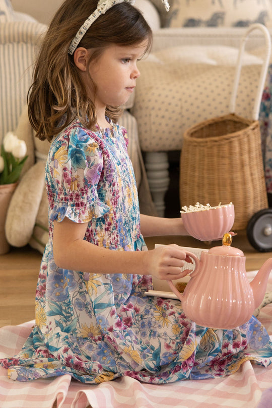 A young girl dressed in the Ivy City Co Mini Cordelia Dress in Pretty Plumeria sits on a checkered blanket, holding a pink teapot and a teacup filled with popcorn as she enjoys an indoor pretend tea party—a charming scene ideal for spring celebrations and treasured family moments.