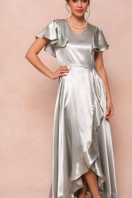 A woman wearing the Ivy City Co Callie Dress in Sage Satin—a shiny sage green satin dress featuring ruffled short sleeves and a ruffled hem—styled with a pearl necklace, stands against a soft beige background.