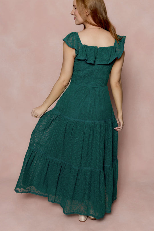 A woman wearing the Alaina Dress in Deep Sea Lace by Ivy City Co stands against a pink background, facing away and slightly turning her head to the side.