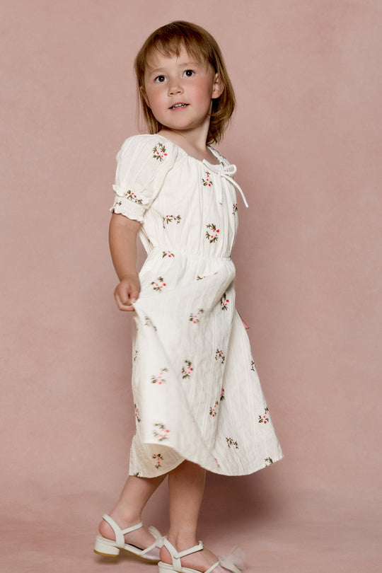 A young child wears the Ivy City Co Mini Jessie Dress in Ditsy Embroidery, paired with white sandals, standing against a soft pink background and gazing gently upward and to the side.