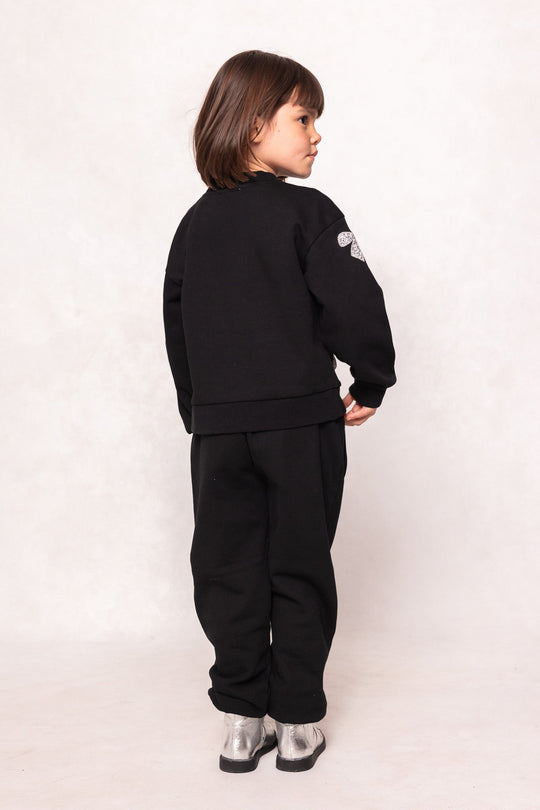 A young child with brown hair stands facing away, wearing a black sweatshirt, Ivy City Co’s Mini Micah Jogger Sweatpants in Black, and silver boots against a plain light background.