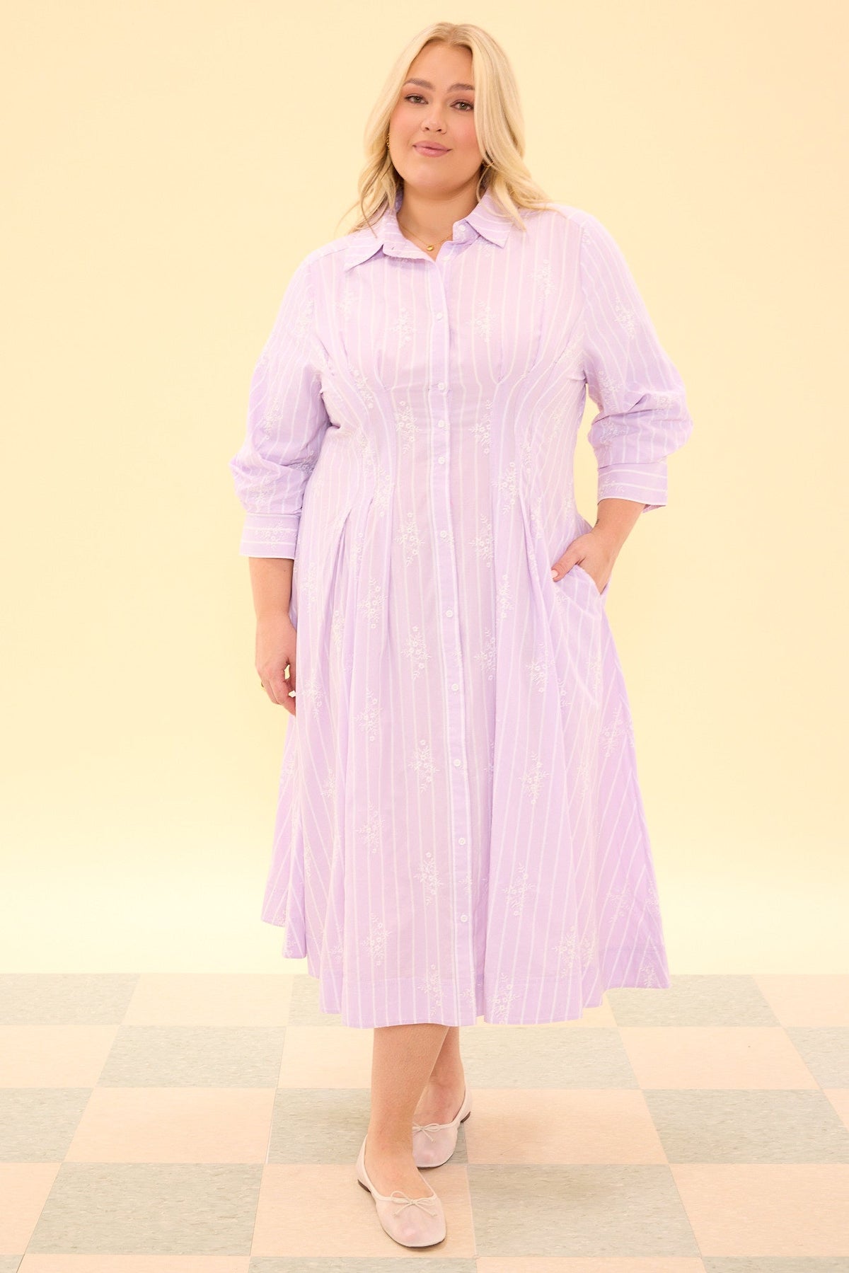 A person wearing the Cameron Dress in Lavender by Ivy City Co—a nursing-friendly, long-sleeve midi shirt dress with a button-down front and light purple pinstripes—stands on a checkered floor, facing forward and smiling slightly. This dress combines comfort and style with its elegant silhouette and subtle lavender stripes.