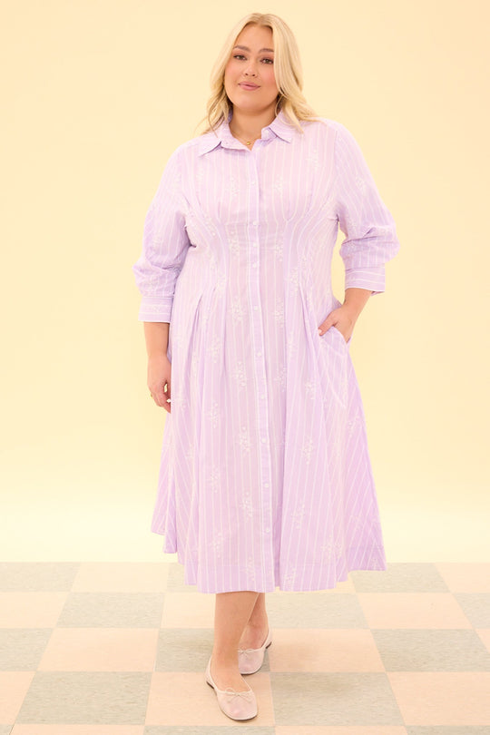 A person wearing the Cameron Dress in Lavender by Ivy City Co—a nursing-friendly, long-sleeve midi shirt dress with a button-down front and light purple pinstripes—stands on a checkered floor, facing forward and smiling slightly. This dress combines comfort and style with its elegant silhouette and subtle lavender stripes.