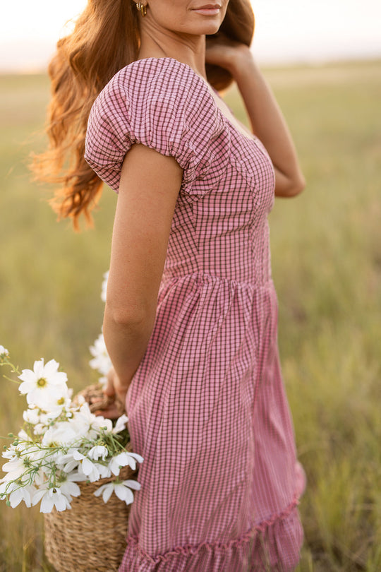 A woman wearing the Ivy City Co Monica Dress in Pink & Brown Gingham stands in a grassy field, holding a woven basket filled with white flowers. The photo is taken from the side with her face partially out of the frame.